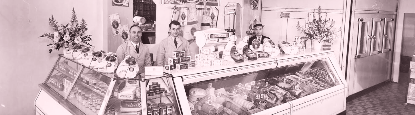 Vintage photo of three men behind a grocery store counter with meats,cheeses and packaged foods