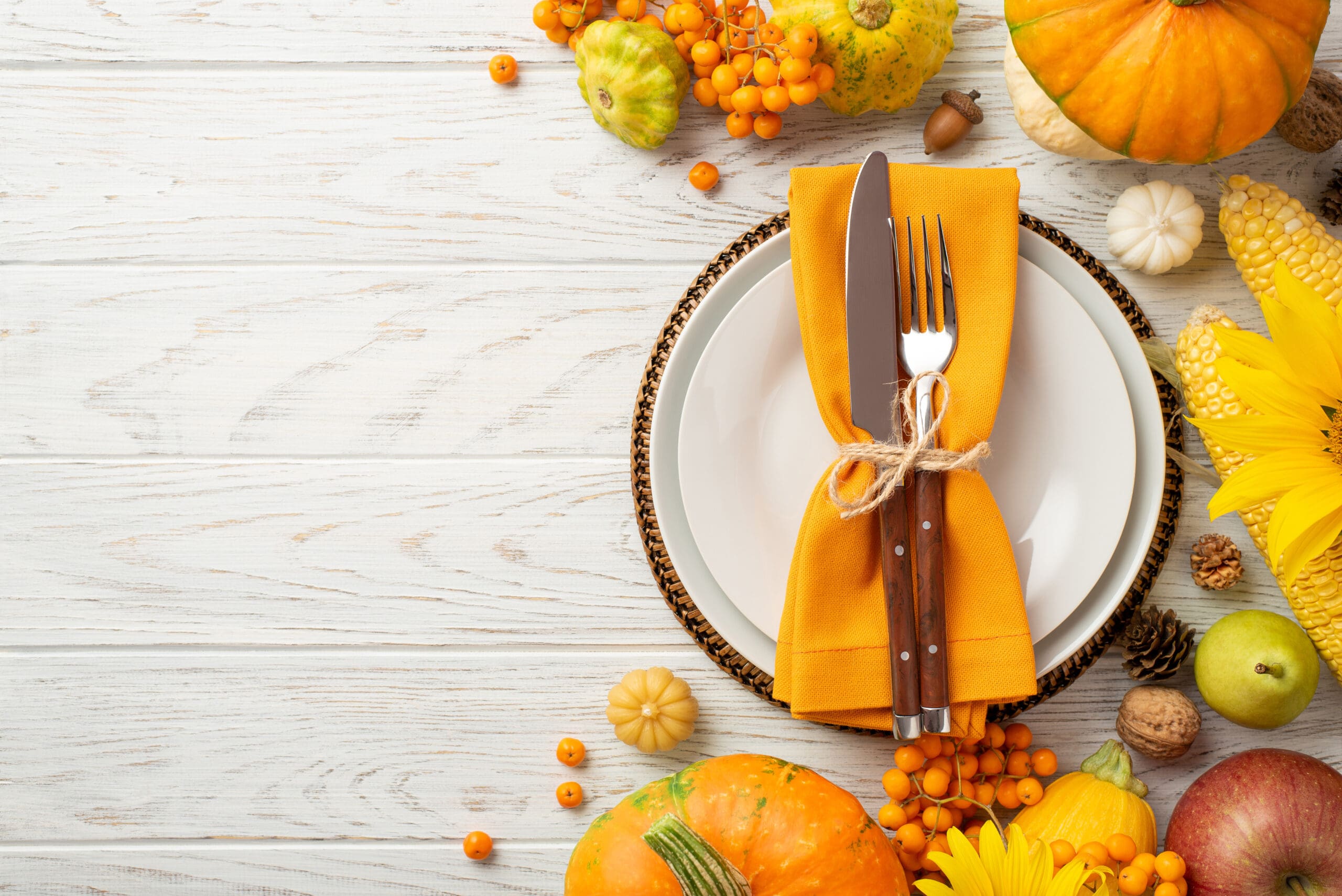 Thanksgiving day concept. Top view photo of plate knife fork napkin raw vegetables pumpkins maize pattypans apple pear walnut acorn pine cones rowan sunflowers isolated white wooden table background