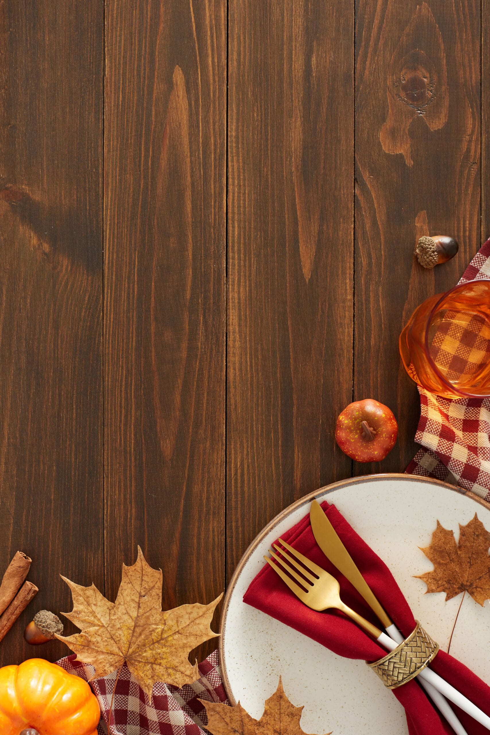 Creating a cozy autumn table. Top view vertical shot of plate, cutlery, napkin, tablecloth, pumpkins, cinnamon sticks, glass, dry leaves on brown wooden table background with space for promo or text